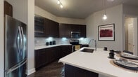 This thoughtfully designed single-floor residence combines contemporary style with functional living across a well-organized layout. The kitchen serves as a standout feature, showcasing rich dark espresso cabinetry with under-cabinet lighting, a classic subway tile backsplash, stainless steel appliances including a French door refrigerator, electric range, and built-in microwave. A large island with a white quartz countertop and undermount sink provides both additional workspace and casual seating, finished with a modern pendant light fixture overhead. Dark hardwood flooring runs throughout the kitchen and living areas, creating a striking contrast against the light-toned walls. The living room offers a comfortable, open feel with sliding glass doors leading to a private outdoor balcony, allowing natural light to fill the space. Multiple bedrooms feature soft carpeting, generous windows with stylish drapery, and well-proportioned layouts suited for full-sized furnishings. The bathrooms are finished with large-format tile flooring, dark wood vanities with white countertops, and framed mirrors. One bathroom includes a glass-enclosed walk-in shower, while the other features convenient access to a walk-in closet. The overall design palette of neutral tones, dark wood accents, and modern fixtures creates a cohesive and refined living environment throughout.
