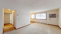 This single-floor unit offers a straightforward, functional layout spread across several well-defined spaces. The living area is generously sized, featuring neutral wall-to-wall carpeting, crisp white walls, and warm reddish-brown baseboards that carry throughout the unit. A large set of double windows along one wall allows abundant natural light to fill the room, offering views of mature trees outside. Baseboard heating units run beneath the windows, and a wall-mounted air conditioning unit provides climate control year-round.

Additional rooms, also carpeted and naturally lit via wood-trimmed windows, offer flexible use as bedrooms or private spaces. Each room maintains the same clean, neutral palette and includes baseboard heating for consistent comfort.

The galley-style kitchen is efficiently arranged with rich oak cabinetry lining both walls, granite-toned countertops, a double-basin sink, and stainless and black appliances including an electric range, microwave, and refrigerator. Durable tile flooring grounds the kitchen, which connects directly to the adjoining rooms for easy flow.

The bathroom features white mosaic tile flooring, full white subway tile surrounds at the tub and shower, a toilet, and a wood-framed vanity with a mirror. The overall ambiance of the unit is clean, neutral, and move-in ready.