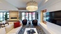 This thoughtfully designed single-floor space combines modern aesthetics with functional elegance across several well-appointed areas. The main lounge serves as the centerpiece, showcasing a striking large-format chandelier with layered crystal elements that draw the eye upward, while polished tile flooring and a patterned area rug define the seating arrangements below. Plush seating, including a white sectional sofa accented with bold red and black pillows and curved swivel chairs, creates an inviting and sophisticated atmosphere. A warm wood-paneled accent wall frames a wall-mounted flat-screen television, adding a refined focal point to the room. Floor-to-ceiling glass sliding doors connect the interior directly to an outdoor patio area, flooding the space with natural light and providing views of an exterior terrace with dining furniture. A secondary lounge area features a white sofa paired with a clear acrylic coffee table, bordered by glass partition walls that maintain an open, airy feel while subtly defining separate zones. The kitchen is sleek and contemporary, outfitted with high-gloss white cabinetry, a black tile backsplash, stainless steel appliances including a refrigerator and dishwasher, and open display shelving adorned with lush greenery. Together, these spaces deliver a polished, modern environment ideal for gathering and entertaining.