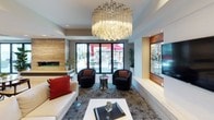 This thoughtfully designed single-floor space combines modern aesthetics with functional elegance across several well-appointed areas. The main lounge serves as the centerpiece, showcasing a striking large-format chandelier with layered crystal elements that draw the eye upward, while polished tile flooring and a patterned area rug define the seating arrangements below. Plush seating, including a white sectional sofa accented with bold red and black pillows and curved swivel chairs, creates an inviting and sophisticated atmosphere. A warm wood-paneled accent wall frames a wall-mounted flat-screen television, adding a refined focal point to the room. Floor-to-ceiling glass sliding doors connect the interior directly to an outdoor patio area, flooding the space with natural light and providing views of an exterior terrace with dining furniture. A secondary lounge area features a white sofa paired with a clear acrylic coffee table, bordered by glass partition walls that maintain an open, airy feel while subtly defining separate zones. The kitchen is sleek and contemporary, outfitted with high-gloss white cabinetry, a black tile backsplash, stainless steel appliances including a refrigerator and dishwasher, and open display shelving adorned with lush greenery. Together, these spaces deliver a polished, modern environment ideal for gathering and entertaining.