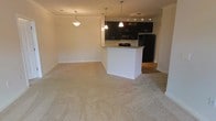 This single-floor space showcases an open-concept layout that seamlessly connects the living area to the kitchen. The living area is finished with plush, neutral-toned carpet that extends throughout, lending a clean and cohesive look to the space. A pendant light fixture anchors the living area, providing warm overhead illumination.

The kitchen is defined by a peninsula with a granite countertop, which serves as both a functional workspace and a natural divider between the living and cooking zones. Dark espresso-toned cabinetry lines the kitchen walls, offering a striking contrast against the light walls and countertops. A set of pendant lights hang above the peninsula, complementing the track lighting mounted on the kitchen ceiling. Black appliances, including a refrigerator and microwave, are integrated into the cabinetry for a uniform, modern aesthetic. The kitchen floor transitions from carpet to a hard surface, further distinguishing the two zones.

Crown molding trims the ceiling throughout, adding a refined architectural detail. The overall ambiance is modern and neutral, with a clean, move-in-ready feel suited for a variety of furnishing styles.