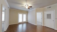 This single-story space offers a clean, well-maintained interior with a thoughtful combination of materials and architectural details. The main living area is finished with rich hardwood flooring that adds warmth and character throughout. Crown molding runs along the perimeter of the ceiling, lending a refined, classic touch to the space. A ceiling fan with an integrated light fixture is centrally positioned, providing both air circulation and ambient lighting.

A pair of white-framed French doors along one wall allows natural light to filter into the room, brightening the neutral, beige-toned walls and creating an inviting atmosphere. The white baseboards and door trim are crisp and well-defined, complementing the overall aesthetic.

An open doorway leads to an adjoining room finished with carpeted flooring, offering a contrast in texture and suggesting a functional separation of living areas. The transition between hardwood and carpet hints at a versatile layout suited for multiple uses. Overall, the space presents a bright, airy ambiance with traditional architectural details, making it a comfortable and adaptable environment.