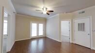 This single-story space offers a clean, well-maintained interior with a thoughtful combination of materials and architectural details. The main living area is finished with rich hardwood flooring that adds warmth and character throughout. Crown molding runs along the perimeter of the ceiling, lending a refined, classic touch to the space. A ceiling fan with an integrated light fixture is centrally positioned, providing both air circulation and ambient lighting.

A pair of white-framed French doors along one wall allows natural light to filter into the room, brightening the neutral, beige-toned walls and creating an inviting atmosphere. The white baseboards and door trim are crisp and well-defined, complementing the overall aesthetic.

An open doorway leads to an adjoining room finished with carpeted flooring, offering a contrast in texture and suggesting a functional separation of living areas. The transition between hardwood and carpet hints at a versatile layout suited for multiple uses. Overall, the space presents a bright, airy ambiance with traditional architectural details, making it a comfortable and adaptable environment.
