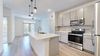 This well-appointed single-floor residence showcases a thoughtfully designed open-concept layout filled with natural light. The kitchen serves as the heart of the home, featuring soft gray cabinetry, a classic tile backsplash, bright white countertops, and a full suite of stainless steel appliances including a refrigerator, electric range, microwave, and dishwasher. A large center island with an undermount sink and pendant lighting above anchors the space and offers additional prep and seating areas. Light wood-tone flooring flows throughout the main living areas, complementing the clean, neutral color palette.

The living spaces are bright and airy, with ceiling fans, large windows, and glass-paneled doors that invite outdoor views and ample natural light. The bedroom areas feature plush carpeting, generous windows overlooking lush greenery, and ceiling fans for added comfort. A spacious walk-in closet provides extensive built-in shelving and dual hanging rods, offering excellent storage capacity.

The bathroom is well-sized, with a wide vanity featuring dual sinks, a framed mirror, and a full tiled soaking tub and shower combination. Light wood flooring carries through this space as well. Throughout the residence, the overall ambiance is clean, contemporary, and move-in ready.