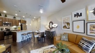 This single-floor residence offers a seamlessly connected open-concept layout that integrates the kitchen, dining, and living areas into one cohesive and functional living space. Dark hardwood floors run throughout, grounding the interior with a warm, sophisticated aesthetic that contrasts beautifully with the crisp white walls and ceilings.

The kitchen is outfitted with rich wood cabinetry, stainless steel appliances, and a light-toned granite or stone countertop breakfast bar with stylish pendant lighting overhead — ideal for casual dining or entertaining. Recessed lighting provides consistent illumination throughout the space.

The dining area accommodates a full table and chair set, complemented by a decorative wine rack console that adds both function and flair. Framed artwork adorns the walls, contributing to the curated, modern ambiance of the home.

The living area is anchored by a warm caramel leather sofa accented with patterned throw pillows, and a ceiling fan overhead ensures year-round comfort. Natural light filters into the space, enhancing its bright, airy feel.

Overall, this residence presents a well-appointed, contemporary interior with thoughtful design details, making it a comfortable and visually appealing living environment.