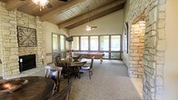 This single-floor recreational space combines rustic charm with functional design, offering an inviting atmosphere for leisure and social gatherings. The room's most striking architectural features are its exposed wooden beam ceilings with tongue-and-groove paneling, which lend a warm, lodge-inspired character to the space. Prominent natural stone columns and accent walls add texture and visual depth throughout, reinforcing the earthy, organic aesthetic.

Large multi-pane windows line the far wall, flooding the interior with generous natural light and providing views of the surrounding greenery outside. Ceiling fans are mounted along the beam ceiling, ensuring comfortable airflow across the space.

The layout is thoughtfully divided to accommodate multiple activities. A dining area near the windows features a sturdy round wooden table surrounded by dark wood chairs, ideal for casual meals or tabletop games. Toward the back of the room, a full-sized billiards table serves as a central recreational focal point. The carpeted flooring throughout provides comfort underfoot and contributes to the room's relaxed, welcoming ambiance.

Overall, this space is well-suited as a community clubhouse or entertainment lounge, offering a harmonious blend of natural materials, recreational amenities, and a comfortable, casual atmosphere.