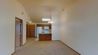 This single-story space features an open-concept layout that combines the kitchen and living area into one cohesive, flowing environment. The vaulted ceilings add a striking sense of height and openness, making the space feel larger than its footprint suggests. A chandelier hangs as a focal point above the living area, complementing the functional fluorescent lighting over the kitchen workspace.

The kitchen is equipped with warm honey-toned wood cabinetry that provides ample storage, along with white appliances including a refrigerator, microwave, and range. A peninsula-style breakfast bar with matching wood cabinetry extends into the living space, offering additional counter space while creating a natural division between the two areas without sacrificing the open feel.

Light-toned flooring spans the living area, while the kitchen features a different hard-surface floor, providing a subtle transition between the two zones. Neutral beige walls throughout create a clean, versatile backdrop. An entry door with a wood finish is visible toward the rear, alongside a hallway that leads to additional areas of the unit. The overall ambiance is functional, understated, and welcoming.