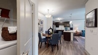 This single-floor residence is thoughtfully designed with an open-concept layout that seamlessly connects the living, dining, and kitchen areas. The living room features wood-style flooring, crown molding, a ceiling fan, and large windows with exterior door access, allowing natural light to fill the space. A sectional seating area anchors the room alongside a glass-top coffee table and coordinated area rug.

The dining area accommodates a round table with upholstered chairs beneath a pendant light fixture, accented by a gallery wall and an indoor plant. It flows directly into the kitchen, which is equipped with dark espresso cabinetry, light quartz-style countertops, stainless steel appliances including a refrigerator, range, and microwave, and a double-basin sink. A kitchen island with pendant lighting provides additional workspace and bar seating.

The bedroom features plush carpeting, a walk-in closet with built-in shelving, and windows that bring in ample natural light with outdoor views. The bathroom includes a full soaking tub with a tile surround in a neutral stone finish and a decorative curtain. Overall, the space presents a cohesive, modern aesthetic with warm tones and functional design throughout.