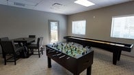 This single-floor space is designed with recreation and social engagement in mind. The game room serves as the centerpiece of the space, offering a variety of entertainment options for guests and visitors. A full-size foosball table takes center stage, while a shuffleboard table lines the wall, providing classic gaming options for friendly competition. Additional seating, including upholstered chairs, is available for those looking to relax or observe the action.

The room features a neutral gray color palette on the walls, complemented by light wood-style flooring that gives the space a clean, modern feel. A drop ceiling with recessed panel lighting ensures the area is well-lit throughout. Natural light filters in through a window fitted with horizontal blinds, adding brightness to the interior. A door with a window provides access to an outdoor area, suggesting a deck or patio beyond, which enhances the sense of openness and connectivity to the outdoors.

Overall, the space strikes a balance between functionality and comfort, making it well-suited as a communal amenity or leisure area. Its straightforward, uncluttered layout allows for easy movement between activities, and the durable finishes suggest the space is built to accommodate regular, active use.