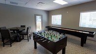 This single-floor space is designed with recreation and social engagement in mind. The game room serves as the centerpiece of the space, offering a variety of entertainment options for guests and visitors. A full-size foosball table takes center stage, while a shuffleboard table lines the wall, providing classic gaming options for friendly competition. Additional seating, including upholstered chairs, is available for those looking to relax or observe the action.

The room features a neutral gray color palette on the walls, complemented by light wood-style flooring that gives the space a clean, modern feel. A drop ceiling with recessed panel lighting ensures the area is well-lit throughout. Natural light filters in through a window fitted with horizontal blinds, adding brightness to the interior. A door with a window provides access to an outdoor area, suggesting a deck or patio beyond, which enhances the sense of openness and connectivity to the outdoors.

Overall, the space strikes a balance between functionality and comfort, making it well-suited as a communal amenity or leisure area. Its straightforward, uncluttered layout allows for easy movement between activities, and the durable finishes suggest the space is built to accommodate regular, active use.