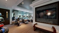 This well-equipped fitness center at 40 Pinehills Dr in Plymouth, Massachusetts, offers a thoughtfully designed workout environment that balances functionality with upscale aesthetics. The space is laid out across a single floor and provides generous room for a diverse array of exercise equipment and activities.

The interior design features a sophisticated palette of soft grays, warm wood-toned flooring, and striped carpeting that delineates different workout zones. Coffered ceilings with recessed lighting and tray-ceiling details in contrasting dark tones lend an elevated, boutique-gym feel to the space. Full-length mirrors along one wall visually expand the room and enhance functionality for users monitoring their form.

Cardio equipment is prominently featured, including multiple treadmills, elliptical machines, and stationary and spin cycles. Strength training is well represented with cable machines, weight benches, medicine balls, and stability balls neatly organized along the perimeter. A built-in media station with wall-mounted flat-screen televisions and integrated cabinetry serves as a central hub, while an additional TV is framed in decorative mosaic tile near a cushioned bench for rest or stretching.

Floor-to-ceiling windows along one wall invite abundant natural light and offer views of the surrounding greenery, creating a refreshing and energizing atmosphere throughout the facility.