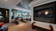 This well-equipped fitness center at 40 Pinehills Dr in Plymouth, Massachusetts, offers a thoughtfully designed workout environment that balances functionality with upscale aesthetics. The space is laid out across a single floor and provides generous room for a diverse array of exercise equipment and activities.

The interior design features a sophisticated palette of soft grays, warm wood-toned flooring, and striped carpeting that delineates different workout zones. Coffered ceilings with recessed lighting and tray-ceiling details in contrasting dark tones lend an elevated, boutique-gym feel to the space. Full-length mirrors along one wall visually expand the room and enhance functionality for users monitoring their form.

Cardio equipment is prominently featured, including multiple treadmills, elliptical machines, and stationary and spin cycles. Strength training is well represented with cable machines, weight benches, medicine balls, and stability balls neatly organized along the perimeter. A built-in media station with wall-mounted flat-screen televisions and integrated cabinetry serves as a central hub, while an additional TV is framed in decorative mosaic tile near a cushioned bench for rest or stretching.

Floor-to-ceiling windows along one wall invite abundant natural light and offer views of the surrounding greenery, creating a refreshing and energizing atmosphere throughout the facility.