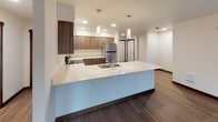 Located at 21044 Viking Way NW in Poulsbo, Washington, this single-floor residence offers a well-designed layout with a clean, modern aesthetic throughout. The open-concept kitchen serves as a central highlight, featuring rich walnut-toned cabinetry extending from base to upper cabinets, light quartz countertops, stainless steel appliances including a refrigerator and range with hood, and a generous island with pendant lighting overhead. The kitchen flows directly into the living area, which is finished with warm wood-look flooring and track lighting, creating a versatile and welcoming space.

The bedrooms are carpeted in neutral tones and feature large windows with views of the surrounding Pacific Northwest woodland, providing ample natural light and a serene atmosphere. Baseboard heaters ensure year-round comfort. The bathrooms are finished with light wood vanities, white quartz countertops, and neutral tile-look flooring; one includes a bathtub and access to a dedicated walk-in closet. A separate laundry room provides added convenience, outfitted with a front-loading washer and walnut-toned upper cabinetry for storage. Dark wood baseboards and door trim carry a consistent design language throughout the unit.