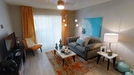 This single-floor residence offers a thoughtfully designed open-concept layout that connects the living, dining, and kitchen areas in a seamless flow. The living room serves as the centerpiece of the home, featuring light hardwood-style flooring, neutral warm-toned walls, and a textured ceiling. A ceiling fan with integrated lighting provides both comfort and illumination, complemented by a large sliding glass door dressed with vibrant orange patterned curtains that flood the space with natural light. The room is anchored by a tufted gray sofa, an abstract area rug in orange and gray tones, and a bold teal wall art piece that adds a pop of color.

The kitchen is efficiently laid out with white cabinetry, dark countertops, stainless steel appliances including a refrigerator and dishwasher, and a dark tile backsplash. A colorful orange runner rug adds a lively touch to the kitchen floor. A compact bar cart near the living area provides a functional and stylish entertaining element.

The bedroom, accessible through a doorway off the main living space, offers a cozy and private retreat. Throughout the residence, the consistent use of warm neutrals, accent colors, and modern furnishings creates a cohesive and inviting ambiance.