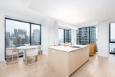 This contemporary high-rise residence is defined by clean lines, a bright white palette, and an abundance of natural light streaming through expansive floor-to-ceiling windows. The open-concept living and dining area serves as the heart of the home, where large black-framed windows frame sweeping panoramic views of the city skyline and waterfront beyond. Light hardwood flooring flows seamlessly throughout, lending warmth to the otherwise crisp, modern aesthetic.

The kitchen is thoughtfully designed with floor-to-ceiling cabinetry in a soft wood-grain finish, a stainless steel range, and a generous island with a clean countertop surface — ideal for both cooking and casual dining.

A long, well-lit corridor connects the living spaces to the private quarters, accented by a large wood-framed mirror that enhances the sense of depth. The primary bedroom is a serene retreat, boasting dramatic corner windows with breathtaking city views and light hardwood floors. Additional bedrooms are equally well-appointed, each featuring ample closet space and a calm, neutral atmosphere. The bathroom offers a clean, minimal design with warm beige tile work and a full soaking tub. Throughout, the residence maintains a cohesive, refined sensibility centered on light, space, and elevated urban living.