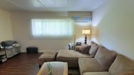 This single-floor space includes a living area designed for comfort and casual use. The room features a large window dressed with vertical blinds, allowing natural light to filter in and offering a view of the greenery outside. A wall-mounted mini-split air conditioning unit is installed above the window, providing efficient climate control throughout the space. The living area is anchored by a spacious L-shaped sectional sofa in a neutral tan tone, complemented by decorative throw pillows, a tufted ottoman, and a rectangular coffee table with a small plant centerpiece. Dark hardwood-style flooring runs throughout, adding warmth and contrast to the light-colored walls. A table lamp with a classic tiered shade provides ambient lighting in the corner. The ceiling features a flat, paneled design. Overall, the space conveys a relaxed, functional atmosphere with practical furnishings and adequate natural light, making it well-suited as a comfortable everyday living environment.