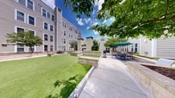 Located in Baltimore's vibrant zip code 21231, this thoughtfully designed outdoor courtyard space is nestled within a multi-story residential complex, offering a welcoming communal retreat for its residents. The courtyard is framed on all sides by the building's neutral-toned horizontal siding and large double-pane windows, creating an enclosed yet open-air atmosphere filled with natural light.

At the heart of the space lies a generous stretch of well-maintained artificial turf, providing a clean, verdant focal point. Surrounding the lawn are raised planters constructed from light-colored stone block, which house a variety of ornamental grasses, ground cover plantings, and mature deciduous trees that offer shade and seasonal greenery. The structured stone planters double as casual seating ledges, enhancing the functional design of the space.

Paved concrete walkways connect the various zones of the courtyard, leading to dedicated outdoor seating and dining areas furnished with tables, chairs, and teal patio umbrellas. Additional lounge seating with blue cushioned furniture is positioned near the raised planters, creating comfortable gathering nooks. Decorative gravel accents and integrated drainage elements reflect the careful attention to landscape detail. A set of bright yellow-framed double doors provides a welcoming entry point into the building, reinforcing the courtyard's role as a central, accessible hub for the community.