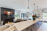 Located at 3400 Peachtree Corners Circle in Peachtree Corners, GA, this single-floor open-plan space seamlessly combines a fully equipped kitchen and a generous living area in a modern, sophisticated design. The living room is anchored by a striking floor-to-ceiling fireplace clad in dark marble with bold white veining, creating a dramatic focal point. A plush tufted sofa, rounded coffee table, and accent chairs are arranged atop a herringbone-patterned light wood floor, which extends throughout the entire space and adds warmth and visual interest. Large black-framed windows and glass-panel doors line multiple walls, flooding the interior with natural light and offering views of the surrounding lush greenery and pool area. The kitchen features a long, sleek island with a white quartz countertop and dark cabinetry, illuminated by a series of modern glass pendant lights. Open wood shelving set against a dark marble backsplash provides both display and storage, while stainless steel appliances and a built-in sink complete the functional layout. A casual dining area with bar-height seating lines the island. The overall ambiance is contemporary and airy, with a refined neutral palette, crisp white walls, and flowing sheer curtains contributing to a bright and welcoming atmosphere.