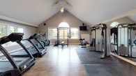 This fitness room is thoughtfully designed to accommodate a full range of workout activities within a single, open-plan space. The architecture is immediately striking, with soaring vaulted ceilings that lend an airy, expansive feel to the room. A prominent arched window, flanked by large rectangular windows, floods the interior with natural light and offers pleasant views of the surrounding greenery, creating an inviting atmosphere for exercise.

The flooring combines warm hardwood with protective rubber gym mats strategically placed beneath equipment zones, balancing aesthetics with functionality. A large wall-mounted mirror on one side visually expands the space while allowing users to monitor their form during workouts.

The cardio zone is well-stocked, featuring multiple treadmills, an elliptical trainer, a recumbent bike, and a rowing machine. The strength training area includes a squat rack with an Olympic barbell and weight plates, a functional cable machine, a tiered dumbbell rack, and various medicine balls. Stability balls and additional free weights round out the equipment selection.

A wall-mounted television provides entertainment, while ceiling-mounted speakers enhance the workout experience. Crown molding and neutral gray walls add a polished, refined finish to the space overall.