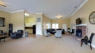 This single-floor space offers an open and functional layout designed for comfort and versatility. The main area is generously proportioned, featuring warm beige walls accented by crisp white crown molding that adds a refined architectural touch throughout. A ceiling fan overhead and multiple windows allow for ample natural light, creating a bright and welcoming ambiance.

The lounge area is furnished with a large sectional sofa and a pair of patterned accent chairs, arranged to encourage comfortable conversation and gathering. The carpeted flooring adds warmth and softness underfoot, while the open floor plan provides flexibility for various uses and configurations.

Adjacent to the main lounge, a kitchenette area is visible and includes white cabinetry, a dark refrigerator, and a countertop workspace. This functional nook is well-suited for light food and beverage preparation, making the space ideal for events or community gatherings.

Additional doors and passageways suggest further utility rooms or access points, contributing to the overall practicality of the layout. The combination of comfortable furnishings, thoughtful architectural detailing, and a well-integrated kitchenette makes this a versatile and inviting space suited for a wide range of social or organizational purposes.