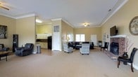 This single-floor space offers an open and functional layout designed for comfort and versatility. The main area is generously proportioned, featuring warm beige walls accented by crisp white crown molding that adds a refined architectural touch throughout. A ceiling fan overhead and multiple windows allow for ample natural light, creating a bright and welcoming ambiance.

The lounge area is furnished with a large sectional sofa and a pair of patterned accent chairs, arranged to encourage comfortable conversation and gathering. The carpeted flooring adds warmth and softness underfoot, while the open floor plan provides flexibility for various uses and configurations.

Adjacent to the main lounge, a kitchenette area is visible and includes white cabinetry, a dark refrigerator, and a countertop workspace. This functional nook is well-suited for light food and beverage preparation, making the space ideal for events or community gatherings.

Additional doors and passageways suggest further utility rooms or access points, contributing to the overall practicality of the layout. The combination of comfortable furnishings, thoughtful architectural detailing, and a well-integrated kitchenette makes this a versatile and inviting space suited for a wide range of social or organizational purposes.