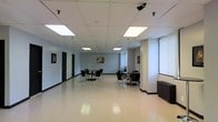 This single-floor commercial space offers a spacious, open-plan interior designed for functionality and flexibility. The layout is clean and expansive, with a polished light-colored floor that reflects the overhead lighting, enhancing the sense of openness throughout the room.

The ceiling features a standard drop-tile system equipped with large recessed fluorescent panels that provide consistent, even illumination across the space. Supplementing these are adjustable track lights mounted near the entrance area, allowing for targeted lighting as needed.

The main area is furnished with multiple small round tables accompanied by dark upholstered chairs, arranged to create distinct informal seating clusters suitable for meetings or collaborative use. Framed artwork is displayed on the neutral gray walls, adding a modest decorative touch to the otherwise utilitarian environment.

Along the far wall, several dark doors lead to private rooms or offices, offering enclosed spaces alongside the open area. A large window fitted with vertical blinds on one side allows for natural light to filter in. The space is well-suited for office use, community gatherings, or small events.