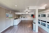 Located at 1501 Harvey Road in College Station, Texas, this community laundry facility offers a clean, functional, and well-organized space designed to serve residents efficiently. The single-floor layout provides a comfortable, open feel with room to move freely between machines and folding areas.

The facility is outfitted with a generous row of top-load washers lining one wall, complemented by a bank of stacked dryers that maximize vertical space along another. All machines are branded and appear to feature digital payment interfaces for convenient use. Built-in countertop shelving runs alongside the washers, providing practical surface space for laundry baskets and supplies.

The interior features wood-look plank flooring throughout, lending a clean and modern aesthetic. Walls are finished with vertical panel detailing in a neutral taupe tone, paired with white crown molding for a polished look. A ceiling fan with a light fixture contributes to air circulation and comfort within the space.

Natural light flows in through multiple double-hung windows along one wall, offering views of the surrounding greenery outside. Recessed lighting and a wall-mounted HVAC unit ensure year-round comfort. A mounted television, security camera, fire extinguisher, and posted instructional signage round out the practical amenities, making this a safe and user-friendly laundry environment.