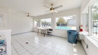 Located at 5 Mooney Rd NE in Fort Walton Beach, Florida, this single-floor community facility serves as a well-equipped laundry and common-use space. The interior is finished with clean white tile flooring throughout, bright white walls, and multiple ceiling fans that promote air circulation and comfort. A wall-mounted mini-split air conditioning unit ensures year-round climate control.

The laundry area is the centerpiece of the space, housing a row of top-load washing machines alongside stacked washer-dryer units, offering ample capacity for residents. Instructional signage is posted on the walls for user convenience. A central island structure provides additional functional support within the laundry zone.

Adjacent to the laundry area, a more open common area benefits from an abundance of natural light streaming through large windows that frame direct views of the outdoor swimming pool and surrounding patio, complete with lounge chairs. This bright, airy section includes a simple table with hairpin legs, offering a casual spot to wait or relax.

A glass-panel entry door provides access to the facility, flanked by windows that further enhance the sense of openness. The overall design is clean, utilitarian, and well-maintained, making this a practical and pleasant shared amenity for the surrounding residential community in Fort Walton Beach.