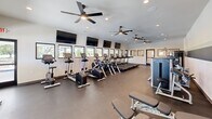 Located at 349 Ridge Rd in Lafayette, Louisiana, this fitness center offers a generously proportioned exercise room designed to support a comprehensive workout experience. The space features a clean, modern aesthetic with crisp white walls accented by subtle horizontal striping, dark-toned flooring, and recessed lighting complemented by multiple ceiling fans that promote airflow and comfort throughout the room.

The cardio zone is prominently featured along one side of the room and includes a row of Life Fitness treadmills, elliptical trainers, and stationary bikes, each positioned facing a bank of wall-mounted flat-screen televisions. A row of horizontal windows runs along the exterior wall, allowing natural light to fill the space and providing views of the surrounding neighborhood.

On the opposite side, a dedicated strength training area houses a selection of Life Fitness selectorized weight machines designed for targeted muscle group exercises. A free weight station features a tiered dumbbell rack stocked with a full range of weights, accompanied by an adjustable workout bench for versatile training options. Additional benches and a cable machine round out the strength section.

The layout is thoughtfully organized to allow comfortable movement between equipment zones. A glass-paneled entry door provides easy access, and the overall ambiance is bright, clean, and motivating — making this fitness center a functional and inviting space for daily exercise.