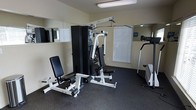 This single-floor fitness room is thoughtfully designed to provide a functional workout environment. The space is anchored by a multi-station strength training machine, offering a variety of resistance-based exercises including cable pulls, leg curls, and pressing movements. An adjustable workout bench accompanies the machine, adding versatility for targeted strength training. A cardio machine, visible along the far wall, rounds out the exercise options available in the space.

The room features durable rubber flooring in a dark speckled finish, ideal for absorbing impact and protecting equipment. Mirrored wall panels are strategically placed to enhance the sense of depth and allow users to monitor their form during workouts. Natural light filters in through a window fitted with horizontal blinds, keeping the space bright while maintaining privacy. The walls are finished in a neutral beige tone with wainscoting detail along the lower portion, giving the room a clean and orderly appearance. Overhead fluorescent lighting ensures consistent illumination throughout. The overall ambiance is practical and focused, making this fitness room a well-appointed space for daily exercise.