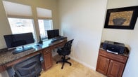 This single-floor office workspace is designed to support productive work with a practical and well-appointed layout. The room centers around a built-in wraparound desk featuring rich granite countertops supported by warm wood cabinetry with drawers and storage below — providing both an elegant aesthetic and substantial workspace. Multiple computer monitors are set up across the desk surface, accommodating more than one user simultaneously, making it well-suited as a shared business center or multi-user workstation area. Ergonomic task chairs accompany the workstations, ensuring comfort during extended use. Natural light fills the room through a set of large windows dressed with clean white roller shades, offering views to the exterior while allowing light control as needed. The light-toned tile flooring complements the neutral, cream-painted walls and white baseboards, creating a clean and professional atmosphere. An additional granite-topped cabinet unit is positioned along the side wall, offering supplementary storage or equipment space. The overall design balances functionality with a polished finish, making it a capable and comfortable environment for business or administrative tasks.