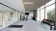 This single-floor fitness center offers a clean, contemporary design that prioritizes both functionality and comfort. The space is defined by its expansive floor-to-ceiling windows along one full wall, flooding the interior with abundant natural light and creating an open, energizing atmosphere for workouts.

The flooring features a patterned modular carpet tile layout in neutral tones of grey and blue, providing a comfortable and durable surface throughout the facility. The ceiling incorporates a mix of acoustic tile panels and recessed linear lighting, ensuring even illumination across the entire space.

The main workout area is thoughtfully organized, with a dedicated row of stationary cycling bikes positioned centrally, ideal for spin sessions. Along the perimeter, a medicine ball rack offers a variety of weighted options for strength and conditioning training. Kettlebells and a balance dome are conveniently positioned near the windowed wall, supporting functional fitness routines. A large wall-mounted flat-screen television provides entertainment and instructional viewing during workouts.

Ceiling-mounted fans offer additional air circulation throughout the space. A glass-paneled entrance door provides a seamless transition into the facility, while the bright, white walls and modern finishes contribute to a clean, professional environment that is both welcoming and motivating.