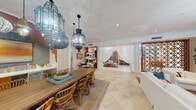 This single-floor amenity space is thoughtfully designed with a cohesive coastal-meets-Moroccan aesthetic that flows seamlessly throughout. The grand entrance lobby greets visitors with soaring tray ceilings, white structural columns, large-pane glass entry doors that invite abundant natural light, and an elegant chandelier overhead. Large-format light tile flooring extends throughout the space, anchoring its bright, airy character.

The open lounge and dining area serves as the heart of the space, centered around a substantial solid-wood dining table paired with rattan chairs accented in teal. Overhead, a dramatic cluster of ornate Moroccan-style pendant lanterns in varying shapes and finishes creates a striking focal point. A built-in wet bar and display niche with warm wood paneling provides a functional and decorative backdrop. French doors offer direct access to an outdoor terrace.

Adjacent to the dining area, a comfortable lounge showcases a large vivid abstract artwork framed in natural wood, complementing the earthy tones of the neutral-hued walls. A second lounge area transitions into a dedicated entertainment room, where crisp white slipcovered sofas face a large flat-screen TV integrated into a floor-to-ceiling built-in shelving unit. A decorative geometric wood screen panel serves as a refined room divider, adding architectural interest. A contemporary starburst chandelier completes the sophisticated yet relaxed ambiance of the space.