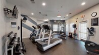 Located at 6959 Castle Ct in Frederick, Maryland, this fitness center is a well-appointed, single-floor facility designed to support a comprehensive workout experience. The space features a modern aesthetic with a striking chevron-patterned accent wall in bold slate blue and white, complemented by recessed lighting and distinctive red pendant lights that lend the room a stylish, energetic ambiance.

The cardio area is equipped with multiple treadmills and stationary bikes, providing ample options for aerobic exercise. A full cable machine and functional trainer anchor the strength-training section, alongside a leg press machine, weight bench, and a rack of colorful kettlebells and medicine balls for varied workout routines. A dumbbell rack rounds out the free weight offerings.

Natural light enters through several windows fitted with white blinds, keeping the space bright and airy. A large wall mirror visually expands the room and allows users to monitor their form during exercise. The durable rubber flooring throughout offers both safety and comfort underfoot.

Practical amenities include a mounted flat-screen TV, a wall clock, a paper towel dispenser, and a waste receptacle. Safety features such as a fire extinguisher and clearly marked exit signage are also present. The overall design balances functionality with contemporary style, making this fitness center a capable and inviting space for residents.