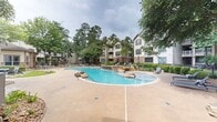 This expansive outdoor amenity area centers around a generously sized freeform swimming pool designed with a resort-style aesthetic. The pool features striking natural rock boulder accents placed along its curved edges and a cascading waterfall element built into a stone retaining wall, creating a serene, naturalistic atmosphere. The pool deck is finished in a warm concrete surface with ample space for lounge seating, and multiple poolside chairs are arranged throughout the area for relaxation.

Adjacent to the pool, a covered outdoor pavilion with a stone-accented base and a pitched roof provides a shaded gathering space, complemented by additional seating and what appears to be an outdoor entertainment setup. Manicured landscaping, including mature pine and oak trees, flowering shrubs, and well-maintained garden beds, frames the entire space, offering privacy and a canopy of natural shade.

The surrounding multi-story residential buildings, constructed with a blend of siding and stone veneer exteriors, feature private balconies that overlook the pool courtyard. Ornamental iron fencing encloses portions of the outdoor space, adding a refined boundary to the grounds. The overall ambiance is lush, tranquil, and well-appointed, reflecting a thoughtfully designed community outdoor living environment.
