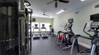 This fitness center offers a thoughtfully arranged workout space designed to accommodate a variety of exercise routines. The room features light green walls that create a clean, energizing atmosphere, complemented by durable rubber flooring ideal for high-impact activities. Natural light filters in through large windows along the far wall, providing a bright and welcoming environment for workouts. Overhead, ceiling fans help maintain comfortable airflow throughout the space. The cardio section is well-stocked with multiple treadmills, a stationary bike, and an elliptical trainer, catering to a range of cardiovascular fitness needs. A stair climber with an integrated display screen adds to the variety of cardio options available. On the strength training side, a multi-functional cable machine is positioned along one wall, offering versatile resistance training possibilities. A stability ball is also present, supporting core and flexibility exercises. Wall-mounted storage racks provide convenient organization for additional accessories. Fluorescent lighting ensures the space remains well-lit at all hours, making it functional regardless of the time of day. Overall, this fitness center combines practical equipment with a comfortable, well-maintained environment.