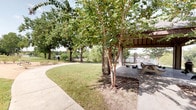 This lakeside outdoor amenity space offers residents a well-appointed recreational environment designed for relaxation and leisure. At its centerpiece is a generously sized swimming pool with crystal-clear water, flanked by an adjoining spa and a broad concrete pool deck lined with lounge chairs and patio umbrellas, providing ample space to unwind in the sun. A covered pavilion structure with rustic dark wood posts and a shaded patio area serves as a transitional space between the interior pool house facility and the outdoor deck, complete with seating for casual gatherings. The pool house building features stucco exterior walls accented by heavy, richly toned timber columns and wooden framed doors, giving the space a warm, lodge-inspired character. Large glass sliding doors connect the sheltered patio to the open pool area, flooding the transition zone with natural light and views of the water. Beyond the pool area, a curving concrete walkway winds through a lush green lawn shaded by a canopy of mature trees, leading toward the lake's edge. Picnic tables are nestled beneath the trees, offering a quiet setting overlooking the water. The surrounding landscape of established trees and open green space creates a peaceful, park-like atmosphere throughout this community amenity area.