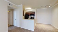 This single-floor residence features a thoughtfully arranged floor plan that maximizes functionality within a compact footprint. The open-concept design connects the kitchen and living area seamlessly, creating a cohesive and accessible living space.

The kitchen is positioned within a partially enclosed nook, defined by a white half-wall that doubles as a breakfast bar or pass-through counter. Rich, dark-stained cabinetry provides ample storage and creates a strong visual contrast against the light-colored walls. Black appliances — including a refrigerator, electric range, and over-the-range microwave — outfit the kitchen with essential amenities. Under-cabinet and overhead fluorescent lighting ensures the kitchen remains well-lit.

The adjacent living area features neutral carpeting and crisp white baseboards and crown molding that lend a clean, finished look throughout. Track lighting on the ceiling adds a modern touch and provides flexible illumination. White interior doors and a consistent neutral color palette throughout contribute to a bright, open ambiance, making the space feel welcoming and move-in ready.