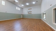 Located at 1900 Westview Blvd in Conroe, TX, this expansive indoor sport court offers a generous, open floor plan designed to accommodate a variety of athletic and recreational pursuits. The space is defined by its impressively tall ceilings, which provide ample vertical clearance for basketball and other court sports, and are fitted with rows of flush fluorescent light fixtures that ensure bright, even illumination throughout. A wall-mounted basketball backboard and hoop are positioned at one end of the court, making the space immediately ready for play. The flooring throughout consists of durable wood-look planks that deliver both aesthetic appeal and a resilient playing surface. The walls feature a two-tone finish — a crisp white upper half and a muted sage green lower half — separated by clean white baseboard trim, lending a polished and purposeful character to the interior. High-set rectangular windows along multiple walls allow natural light to filter in while maintaining privacy and wall space. A pass-through window on one wall connects to an adjacent room, suggesting additional support facilities nearby. The space is accessed via a dedicated door and is well-ventilated through ceiling-mounted HVAC vents. Overall, this court presents a clean, functional, and well-maintained environment suited for basketball, fitness training, or multi-purpose recreational use.