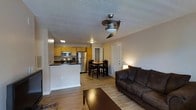 Located at 2800 Ellsworth St in Greenville, NC, this single-floor unit offers a practical and well-organized open-concept layout that connects the living, dining, and kitchen areas seamlessly. Light wood-tone laminate flooring runs throughout the main living space, contributing to a warm and cohesive aesthetic.

The living area is anchored by a plush dark sofa and a wood coffee table, with a ceiling fan and integrated light fixture overhead providing both comfort and illumination. Neutral-toned walls keep the space feeling bright and versatile.

The kitchen is positioned toward the back of the open floor plan and features honey-toned wood cabinetry, a dark countertop peninsula, stainless steel appliances including a refrigerator, microwave, and range, and a skylight that introduces additional natural light. The peninsula provides a natural divider between the kitchen and the rest of the living space.

Between the kitchen and living room, a dining area comfortably accommodates a dark wood table with matching chairs. A hallway door visible from the main room leads to additional spaces within the unit. Overall, the layout is functional and efficient, making good use of the available floor space.