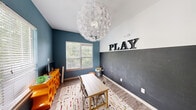 Located at 1100 Townsend Boulevard in Fredericksburg, Virginia, this single-room space is thoughtfully designed as a dedicated play and activity area. The room makes an immediate visual impression with its bold two-tone wall treatment — a rich teal-blue upper half transitions to a full chalkboard surface below, offering both a striking aesthetic and a functional, interactive writing surface. Bold marquee-style letters spelling "PLAY" are mounted above the chalkboard, reinforcing the room's creative purpose.

The space is anchored by a striking dandelion-inspired chandelier that serves as a dramatic focal point overhead, adding an artistic, whimsical touch. Wood-look flooring runs throughout, complemented by a colorful area rug that adds warmth and texture underfoot. Multiple windows with horizontal blinds allow abundant natural light to fill the room, offering views of surrounding greenery.

The room connects to an adjacent living and dining area through a set of elegant French doors with glass pane inserts, providing both privacy and an open, connected feel when desired. The overall ambiance is energetic yet functional — a well-lit, imaginatively styled room well-suited for creativity, learning, and play.