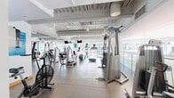 Located on Wilshire Boulevard in Beverly Hills, this fitness center offers a well-appointed, single-floor workout facility designed to accommodate a full range of exercise needs. The space is bright and open, with large windows along one side allowing abundant natural light to fill the room and providing views of the surrounding area.

The layout is thoughtfully organized to maximize functionality. A dedicated cardio zone features multiple treadmills, elliptical trainers, upright and recumbent bikes, and an air resistance bike, all positioned to take advantage of the natural light from the window-lined wall. Mounted flat-screen monitors are installed throughout the ceiling area for entertainment during workouts.

The strength-training section is equally well-equipped, housing a variety of cable machines, resistance machines, and multi-functional weight stations by Precor and similar commercial-grade brands. A functional training area includes TRX suspension trainers and a cable pull system, offering versatility for bodyweight and functional movement exercises.

The flooring consists of large-format rubber tile throughout, providing durability and cushioning. The exposed ceiling features painted structural elements and visible ductwork, lending the space an industrial-modern aesthetic. Floor-to-ceiling mirrors are positioned strategically to enhance the sense of space and support proper form during exercise. The overall ambiance is clean, professional, and well-maintained.
