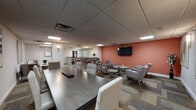This single-floor space is thoughtfully designed to serve multiple functions, combining a comfortable lounge, a bar-style social area, a conference zone, and a fully equipped kitchen within one cohesive layout. The interior is finished with a drop panel ceiling fitted with recessed fluorescent lighting throughout, providing consistent, even illumination across all areas.

The lounge area is anchored by a warm terracotta accent wall, against which a wall-mounted flat-screen television is prominently positioned. Comfortable gray upholstered armchairs are arranged around low, round wooden coffee tables, creating an inviting setting for informal gatherings or relaxed seating. Patterned carpet tiles cover the lounge and conference zones, adding visual texture to the flooring.

A central bar-height counter serves as a focal point between the lounge and the rest of the space. Clad in stacked stone, the counter features granite-style surfaces and is illuminated by decorative pendant lights, with bar stools providing casual seating. Behind it, a mosaic tile backsplash and dark wood cabinetry add refined detail.

A conference table with upholstered seating occupies an adjacent area, suited for structured meetings. The fully equipped kitchen includes dark wood cabinetry, granite countertops, stainless steel appliances — including a gas range, microwave, and refrigerator — a utility sink, and large-format tile flooring.
