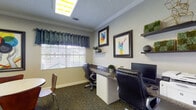 This business center offers a thoughtfully designed, single-floor workspace that blends functionality with a welcoming aesthetic. The main workstation area features a long, shared desk with a dark granite-style surface accommodating two ergonomic leather office chairs and computer monitors, making it ideal for productivity. A window dressed with a decorative dark valance and white blinds allows natural light to filter in, brightening the carpeted workspace. Decorative floating shelves and colorful framed artwork — including vivid animal-themed prints — add personality and visual interest to the neutral-toned walls, which are finished with elegant crown molding throughout. Adjacent to the workstation area is a relaxed lounge and meeting zone, furnished with a round white table, mixed seating including modern side chairs and a tufted ottoman, and a pair of plush black armchairs with built-in side tables. Bold, large-scale framed artwork depicting a cycling fox anchors this lounge area with an energetic, artistic flair. A circular black mirror above the ottoman adds a stylish accent. The space is finished with patterned carpet flooring and bright overhead lighting, creating a comfortable and inspiring environment for work or informal meetings.
