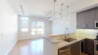 This well-appointed single-floor unit offers a clean, contemporary design with an open-concept layout that seamlessly connects the kitchen and living areas. Light hardwood flooring runs throughout the main living space, reflecting natural light and contributing to the bright, airy atmosphere. Large windows in the living area frame outdoor views of palm trees and the surrounding cityscape, while a ceiling fan ensures comfortable airflow.

The kitchen is a standout feature, equipped with full stainless steel appliances including a gas range, built-in microwave, French door refrigerator, and dishwasher. Rich, dark-toned cabinetry extends to the ceiling, providing ample storage, and is complemented by granite countertops with subtle light veining. A peninsula with a double undermount sink and a professional-style pull-down faucet serves as both a functional workspace and a natural divider between the kitchen and living area. Pendant lighting above the peninsula adds a refined finishing touch.

The bathroom features tile flooring, a soaking tub with textured wave-patterned tile surround, and a separate glass-enclosed shower. The overall design palette is neutral and modern, with white walls, clean lines, and tasteful finishes throughout, creating a cohesive and move-in-ready living environment.