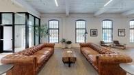 Located at 240 4th St in Inman, South Carolina, this single-floor space is a masterfully converted industrial building that blends historic character with modern functionality. The interior is defined by its whitewashed exposed brick walls, high wood-planked ceilings with industrial steel beams, and polished concrete floors that reflect the open, airy atmosphere throughout.

Tall, arched multi-pane windows line the walls, flooding the expansive open floor plan with generous natural light and offering views of the surrounding streetscape. Structural columns punctuate the interior, reinforcing the building's industrial heritage while adding architectural rhythm to the layout.

The main gathering area is furnished with classic tufted leather Chesterfield sofas, leather lounge chairs, and mixed seating arrangements around café-style tables, creating an inviting and versatile environment. A ping-pong table serves as a recreational centerpiece, complemented by lush potted trees and curated wall art that add warmth and personality. A kitchenette area, accented with bold geometric wallpaper, includes a full-size refrigerator, microwave, and counter space for convenience. Wall-mounted shelving with decorative accents adds a thoughtful finishing touch. This space is well-suited for events, collaborative work, or community gatherings.