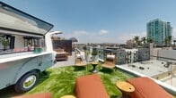 Located at 201 The Promenade N in Long Beach, California, this rooftop terrace offers a well-appointed outdoor amenity space with sweeping views of the surrounding downtown skyline and distant mountains. The terrace is finished with lush artificial turf throughout, creating a clean, park-like atmosphere open to the Southern California sky.

The space is thoughtfully divided into several distinct activity zones. An outdoor cinema area features a large projection screen mounted against a warm wood-clad wall, accompanied by clusters of oversized bean bag seating and accent tables—ideal for communal gatherings and screenings. String lights overhead add a festive ambiance as day transitions to evening.

A separate lounge area is furnished with wicker and rattan chairs, chaise lounges, and side tables, providing a relaxed setting for socializing. A vintage-style trailer serves as a bar or service station, adding a playful, eclectic design element.

The entertainment zone includes a foosball table, rocking lounge chairs with colorful striped cushions, and a central circular fire pit surrounded by additional seating. Glass railings along the perimeter preserve unobstructed panoramic views of Long Beach's urban landscape, making this rooftop a dynamic and versatile outdoor destination.