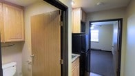 This single-floor space is designed for practical, efficient living. The kitchenette serves as the heart of the unit, equipped with black appliances including a full-size refrigerator and microwave, complemented by light wood cabinetry that extends to the upper walls for generous storage. A sink with a granite-style countertop adds a functional work surface, while dark trim accents throughout provide a clean visual contrast against the lighter cabinetry and walls. A solid wood interior door, finished in a warm natural tone with black hardware, separates areas and contributes to the unit's straightforward, no-frills aesthetic. Adjacent to the kitchenette, a carpeted room is visible, offering a softer, more comfortable space suitable for living or sleeping. This room benefits from natural light provided by a window and is fitted with a ceiling fan for air circulation. The overall layout is compact yet well-organized, making efficient use of the available floor area. The consistent use of dark trim, black appliances, and neutral wall tones creates a cohesive, understated interior that prioritizes functionality and ease of maintenance throughout the space.