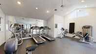 This dedicated fitness center offers a comprehensive workout environment designed to accommodate a wide range of exercise needs. The space features vaulted ceilings that lend an open, airy feel, while large windows fitted with plantation shutters allow generous natural light to fill the room throughout the day. The flooring throughout is durable dark rubber, ideal for high-traffic fitness use.

The cardio zone is well-appointed with multiple treadmills and a recumbent stationary bike, complemented by a large wall-mounted mirror that visually expands the space and allows users to monitor their form. A ceiling fan ensures comfortable airflow during workouts.

The strength training area is equally well-stocked, featuring a full rack of hex dumbbells in graduated weights, a set of medicine balls on a vertical rack, and a Precor cable machine for versatile resistance training. Additional strength equipment, including a weight bench and a functional trainer, round out the selection.

Near the entrance, a built-in shelving unit with a glass-paned door provides organized storage for equipment accessories and supplies. A glass exterior door connects the fitness center to the outdoor surroundings, offering convenient access and additional ventilation. The overall layout is thoughtfully arranged to maximize usable floor space while maintaining clear circulation paths throughout the facility.