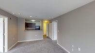This single-floor space is designed with an open-concept layout that connects the living area to the kitchen. The living space is finished with wall-to-wall carpeting in a neutral gray tone, complemented by light gray walls and white baseboards that create a clean, cohesive aesthetic throughout.

The kitchen is positioned at the far end of the room and features a raised breakfast bar pass-through, providing both a visual separation and a functional counter surface. Dark espresso-toned cabinetry lines the kitchen walls, and recessed lighting illuminates the workspace. A microwave is visible among the appliances, and the kitchen benefits from its own dedicated overhead lighting.

From the living area, a hallway leads toward additional rooms, with a warm-toned interior visible through an open doorway suggesting a well-lit adjoining space. Two white bi-fold or hinged doors along the adjacent wall indicate ample closet or storage capacity. Recessed ceiling lights are distributed throughout, ensuring consistent illumination. The overall design is functional and understated, offering a versatile, move-in-ready environment.